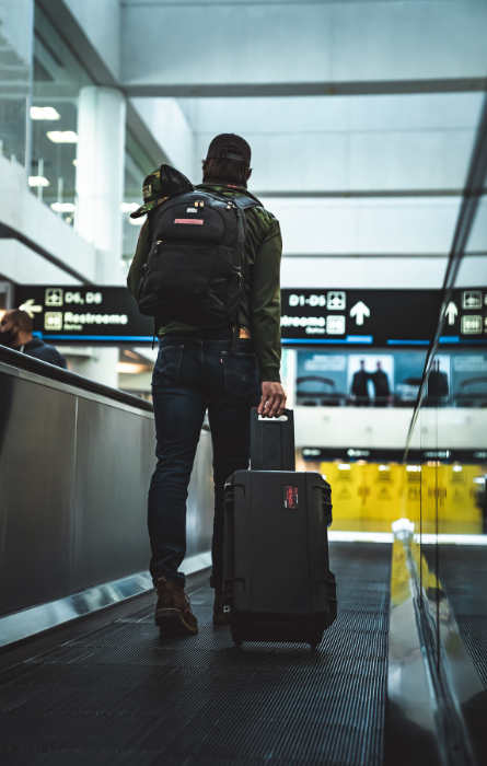 Reiziger op luchthaven met zwarte SKB soft case op wielen en rugzak, geschikt voor het veilig vervoeren van apparatuur tijdens reizen.