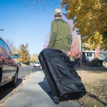 SKB nylon tas met wielen, voortgetrokken door een persoon op een stoep in een woonwijk met geparkeerde auto’s en herfstbomen.